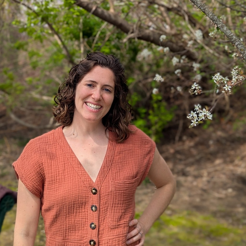 White woman with brown curly hair smiling in a red jumpsuit, in front of a tree with white flowers